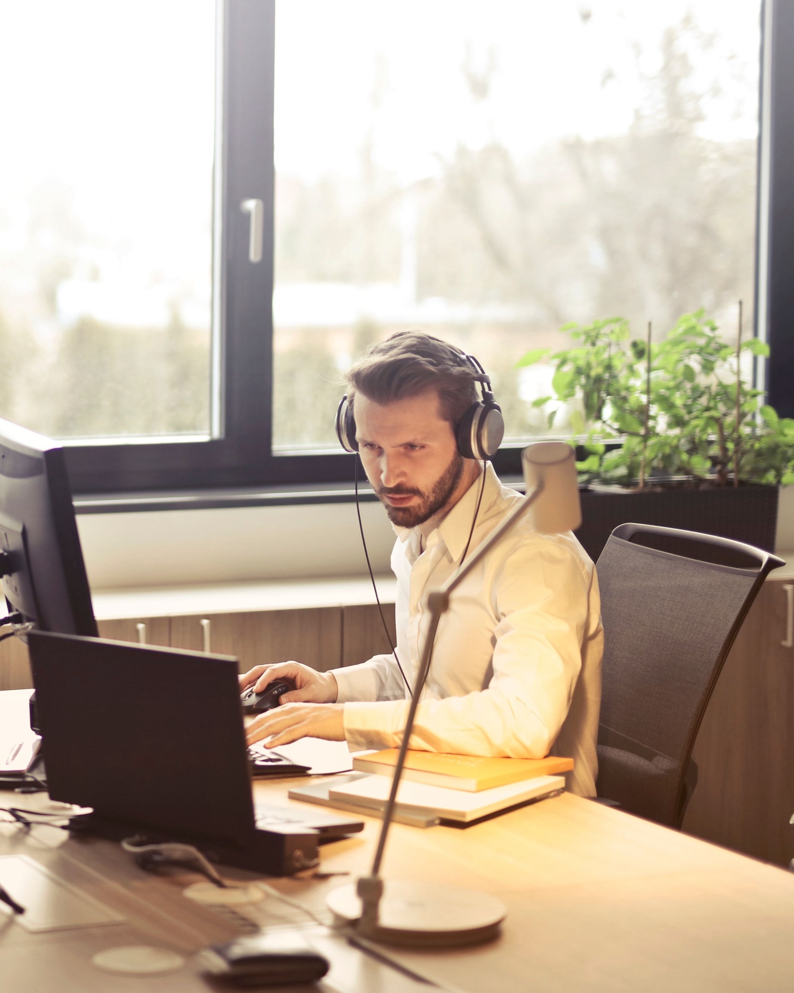 A man wearing headphones working at a desk by a window